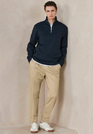 Young man standing with hands in pockets, wearing a navy quarter-zip sweater, beige trousers, and white sneakers against a neutral background.