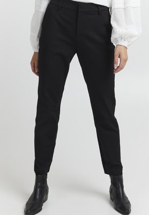 Femme portant un pantalon noir cintré, un chemisier blanc à manches longues bouffantes et des bottines noires, debout devant un fond uni.