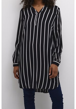 Femme portant une tunique noire à manches longues avec des rayures blanches verticales et un pantalon bleu foncé, debout devant un fond clair uni.