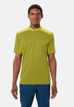 Mann trägt ein limettengrünes kurzärmeliges Hemd mit Frontreißverschluss und dunkelblaue Hose, steht vor einem einfarbigen Hintergrund.