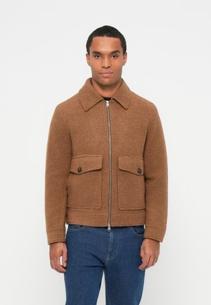 Brown wool jacket with a pointed collar, zip front, two chest pockets, and a fitted design, paired with blue denim jeans.