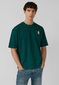 Young man with curly hair wearing teal T-shirt featuring a small Snoopy with Santa hat holding a bell on left chest, and light blue jeans.