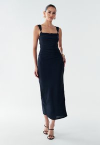 Juoda midi suknelė su prigludusiu liemeniu, kvadratine iškirpte ir surangytomis detalėmis. Turi platų petnešėlių su metalinėmis detalėmis, lygų audinį ir subtilų blizgesį.