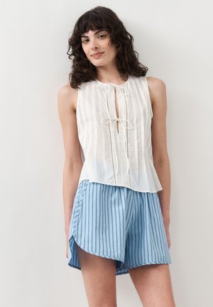 Γυναίκα με λευκό αμάνικο μπλουζάκι με δεσίματα μπροστά και ανοιχτό μπλε ριγέ σορτς, στέκεται μπροστά σε απλό ανοιχτό φόντο.