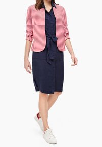 Rood-wit gestreepte jas over een marineblauwe jurk met een strikceintuur, gecombineerd met witte sneakers met een rode accenten.