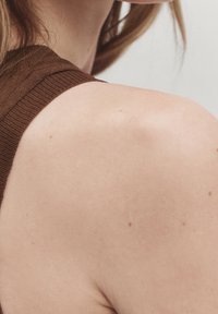 Ruskea neulottu olkaimellinen toppi, jossa on ribbineulostekstuuri, paljastaen sileää ihoa ja hienovaraisia pisamia vaaleaa taustaa vasten.