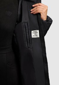 Black puffer jacket with a quilted inner lining, featuring a label reading “A warm hug made out of cloth” and a side pocket.