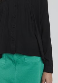 Maglietta nera a maniche lunghe con una finitura testurizzata e chiusura a bottoni, abbinata a pantaloni verdi con tasche e un tessuto liscio.
