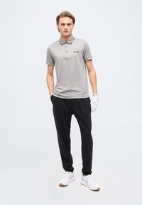 Νέος άνδρας όρθιος με τα χέρια στις τσέπες, φορώντας ανοιχτό γκρι πόλο BOSS, μαύρο παντελόνι, λευκό γάντι στο δεξί χέρι και λευκά αθλητικά παπούτσια.