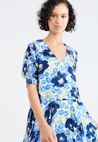 Blauwe bloemenjurk met korte pofmouwen, V-hals en een geraffineerde rok, met een mix van donkere en lichte blauwe bloemen op een witte achtergrond.