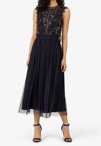 Donkerblauwe jurk met kanten lijfje en geschulpte randen, voorzien van een geplooide tule rok. Gecombineerd met bijpassende blauwe sandalen met bandjes.