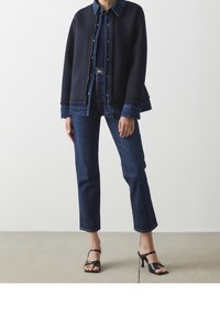 Marinblå sweatshirtjacka över en denimskjorta, tillsammans med mörkblå raka jeans och svarta klacksandaler. Slät textur, avslappnad design.