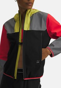 Schwarze, rote, graue und gelbe Zip-Jacke mit Kapuze; verfügt über zwei Reißverschluss-Taschen und einen strukturierten Stoff. Mit roten Akzenten versehen.