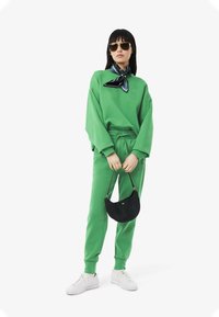Grön sweatshirt och matchande sweatpants i mjukt material, kombinerat med en svart texturerad väska och vita sneakers. Scarfen ger en accentdetalj.