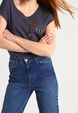 Femme portant un t-shirt gris foncé orné d'écussons de style militaire et un jean bleu taille haute, bras croisés, cheveux partiellement visibles flottant vers la droite.