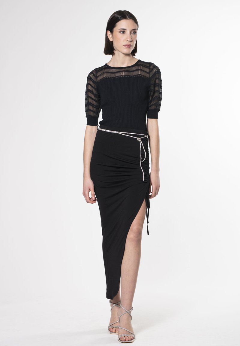 Schwarzer, kurzer Pullover mit durchsichtigen gestreiften Ärmeln, kombiniert mit einem schwarzen, gerafften Rock mit seitlichem Schlitz und gebundenem Akzent. Silberne, strappy High Heels.