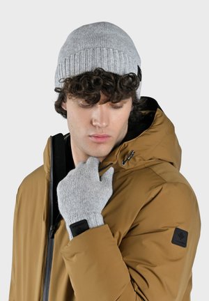 Ung mand iført en beige vinterjakke med hætte, grå strikket hue og matchende grå handsker, som justerer kraven på jakken, neutral baggrund.