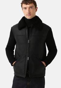 Zwart leren vest met een gestructureerde zwarte gebreide mouw, ritssluiting aan de voorkant en twee schuin geplaatste zakken. Voorzien van een zachte, bontachtige kraag.