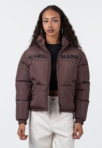 Rudas pūkų striukė su trumpu dizainu, turinti elastingas rankoves, aukštą apykaklę ir išsiuvinėtą "KARL KANI" logotipą priekyje.