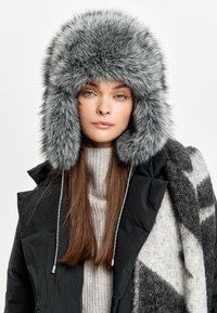 Șapcă gri din blană artificială cu țesături mari pentru urechi, textură moale și volum generos. Se asortează cu o haină neagră și un pulover tricotat gri.