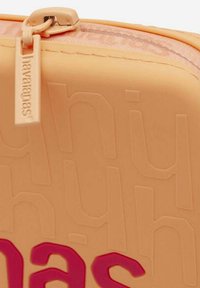 Custodia cosmetica in silicone arancione con logo in rilievo, dettagli rosa, chiusura a zip e superficie texturizzata. Forma rettangolare compatta.