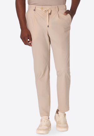 Uomo che indossa pantaloni beige slim fit con vita regolabile a laccio e sneakers beige, in piedi con una mano in tasca davanti a uno sfondo semplice.