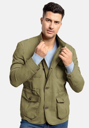 Chaqueta verde oliva con cierre de botones, dos bolsillos frontales y un acabado texturizado. Llevada sobre un suéter de color azul claro.