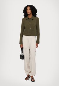 Λαδί κοντό μπουφάν με ασημί κούμπωμα, σε συνδυασμό με χαλαρές κρέμ παν trousers και μαύρη τσάντα χειρός. Μαύρες σαγιονάρες με λουριά ολοκληρώνουν την εμφάνιση.