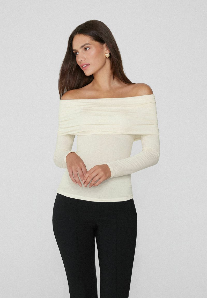 Cremefarbene, schulterfreie Bluse mit langen Ärmeln und geraffter Textur. Kombiniert mit schwarzen Hosen, die eine figurbetonte Silhouette und glattes Material präsentieren.