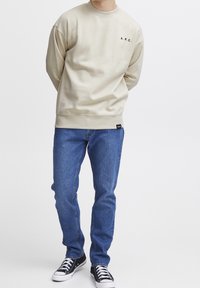 Mężczyzna w beżowej bluzie z napisem "A.R.C.", niebieskich dżinsach i czarnych trampkach stoi na tle białym.