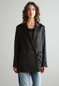 Svart dobbeltspent blazer med strukturert finish, to frontlommer, klaffelapper og avslappet passform, båret over en svart topp og lyse denimjeans.