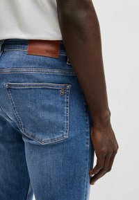Jeans azul oscuro de corte slim, con un parche de cuero con el logo de BOSS en la parte trasera, dos bolsillos traseros con detalles de costura y una textura suave.