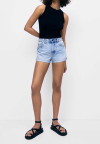 Top sin mangas de canalé negro combinado con pantalones cortos de mezclilla azul claro y sandalias negras de tiras. Los pantalones cortos tienen un dobladillo enrollado y un diseño clásico de cinco bolsillos.