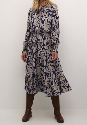 Kobieta ubrana w granatową sukienkę z beżowym wzorem paisley, długimi rękawami, marszczoną talią i brązowymi wysokimi butami, na gładkim tle.