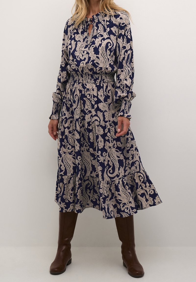 Femme portant une robe bleu marine avec un imprimé beige paisley, manches longues, taille smockée et bottes marron montant jusqu'aux genoux sur un fond uni.