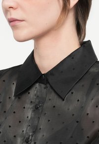 Detailný záber osoby v priehľadnej čiernej blúzke s malými srdcovými vzormi a pevným čiernym špicatým golierom.