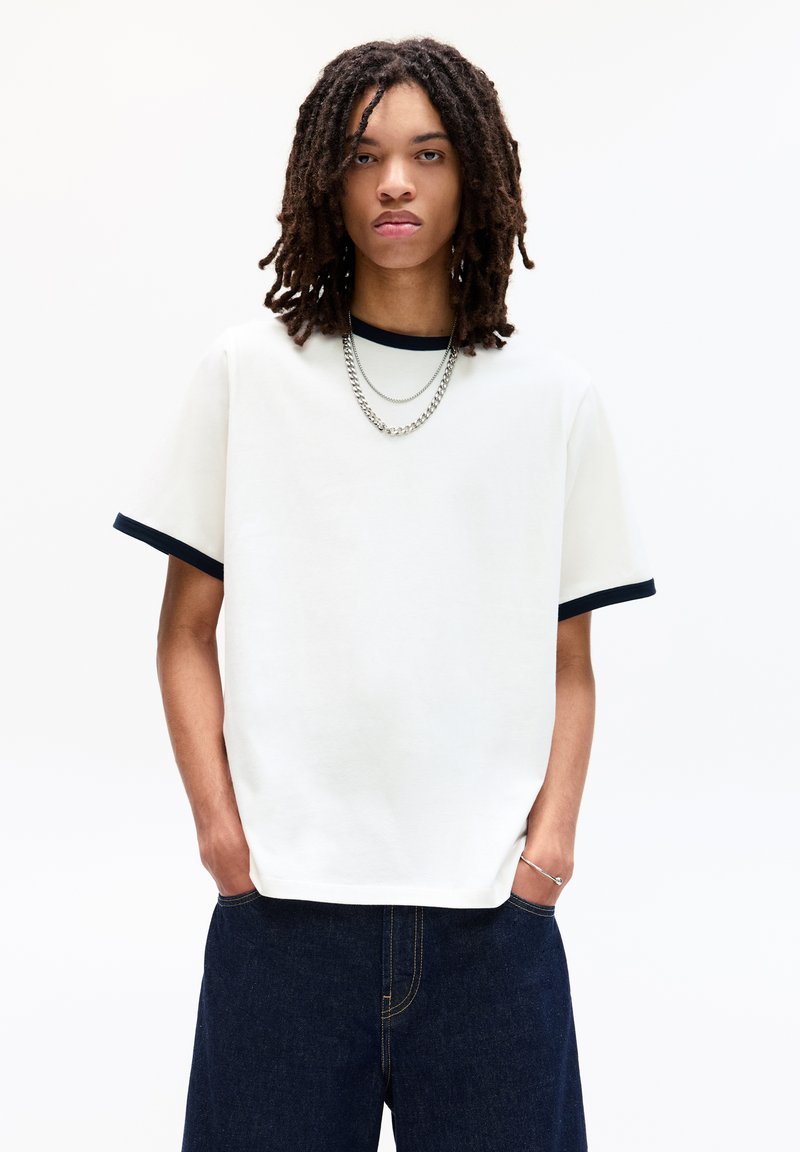 Hvid kortærmet t-shirt med marineblå kant, afslappet pasform, lavet af bomuldsstof. Bæres sammen med mørke denimbukser og lag af sølvkæder.
