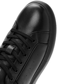 Zapatilla negra con una parte superior de suave cuero sintético, puntera reforzada, cordones negros planos y detalles perforados en el lateral.
