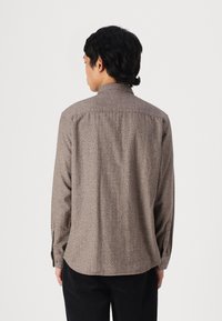 Μακρυμάνικο πουκάμισο σε καφέ χρώμα με υφασμάτινο σχέδιο, διαθέτοντας γιακά με κουμπιά και τσέπες στο στήθος, συνδυασμένο με μαύρο παντελόνι.