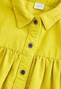 Tissu en velours côtelé jaune avec col et trois boutons noirs sur le devant froncé d'une chemise, affichant l'étiquette de la marque NEXT à l'intérieur du col.