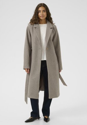 Long beige coat with a lapel collar, single button closure, and side pockets, paired with dark jeans and black pointed flats.
