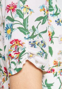 Blusa floral con margaritas y lirios coloridos sobre un fondo blanco. Presenta mangas fruncidas y una tela suave y texturizada.