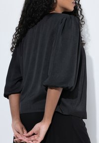 Blusa nera con maniche a sbuffo, tessuto morbido e vestibilità rilassata. La vista da dietro mostra un leggero drappeggio sulle spalle e un fondo dritto.
