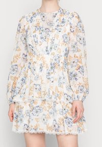 Blommig klänning med långa puffärmar i genomskinlig vit tyg. Har blåa och gula blommor, en lager-på-lager kjol och spetsdetaljer.