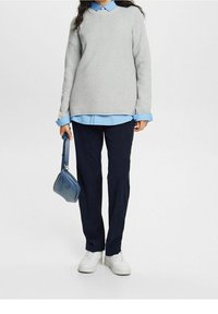 Lichtgrijze textuurtrui over een lichtblauw collshirt, gecombineerd met marineblauwe broek en witte sneakers. Houdt een kleine blauwe handtas vast.