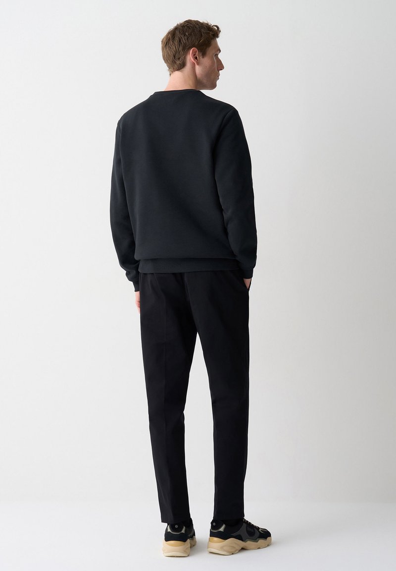 Man met de rug naar de camera, gekleed in een zwart sweatshirt, zwarte broek en zwarte sneakers met beige zolen, tegen een effen witte achtergrond.