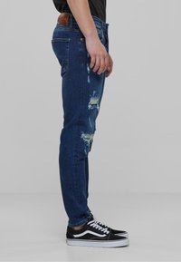 Jeans en denim bleu avec des détails usés à l'avant. Conception ajustée, avec un patch en cuir marron à l'arrière et associés à des baskets noires.