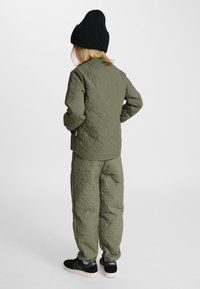 Dziecko stojące tyłem, ubrane w oliwkową pikowaną kurtkę i spodnie, czarną czapkę oraz czarne trampki na jednolitym tle.