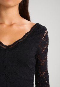 Frau, die ein schwarzes Spitzenoberteil mit V-Ausschnitt und durchsichtigen, floral gemusterten Spitzenärmeln trägt, von den Schultern bis zur Mitte des Oberkörpers vor einem hellen Hintergrund gezeigt.