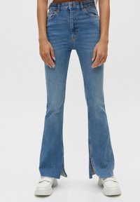 Flared blue denim jeans with a high waist, featuring side slits at the hem, stitched accents, and a smooth texture. Paired with white sneakers.